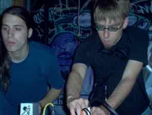 Two performers behind gear at a dark warehouse event, graffiti on the wall behind them