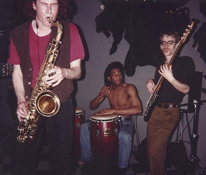 A trio playing in a dark room: a saxophonist, a conga player, and John von Seggern on bass guitar