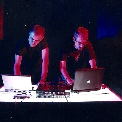 Two performers lit red and blue, bent over laptops and a mixer on a white table