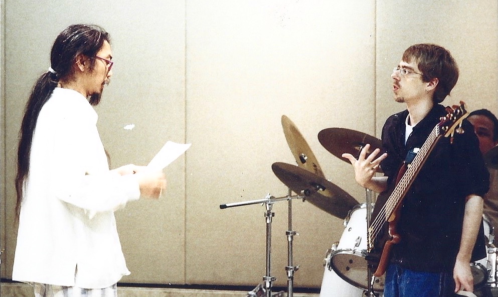 Kung Chi Shing reading from a score while John von Seggern, bass guitar in hand, talks back across a rehearsal room