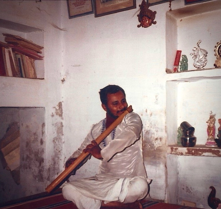 R.J. Srinivasan seated on the floor of a whitewashed room, playing a long bamboo flute, a bookshelf and small shrine niches behind him